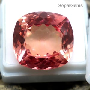 Puede incluir: Una gema facetada de color melocotón en una vitrina blanca. La gema tiene un corte cojín cuadrado y está colocada sobre un fondo texturizado gris-marrón. El texto "SepalGems" es visible.