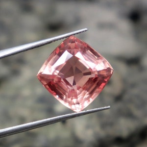 May include: A pink gemstone with a square cushion cut. The gemstone is held by a pair of tweezers.