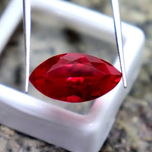 May include: A bright red marquise-shaped gemstone held by tweezers. The gemstone is in a white square display box.
