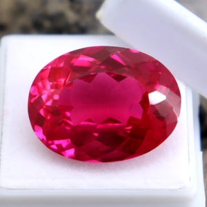 May include: A faceted, oval-shaped, vibrant red gemstone. The gem is set in a white plastic case, with the lid partially open. The gemstone's cut reflects light, creating a sparkling effect. The background is a blurred mix of neutral tones.