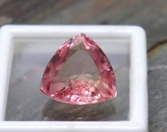 May include: A pink gemstone with a triangular cut, displayed in a white display box.