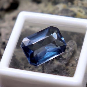 Puede incluir: Una piedra preciosa azul oscuro con un corte rectangular, exhibida en una caja blanca con una tapa transparente.