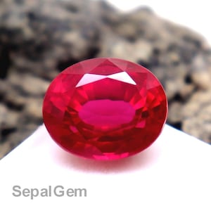 Op de afbeelding: Een ovaalvormige, gefacetteerde robijn edelsteen, met een levendige rode kleur. De edelsteen ligt op een wit oppervlak, met een getextureerde, grijs-bruine rotsformatie op de achtergrond. De tekst "SepalGem" is zichtbaar.
