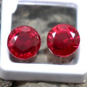 May include: Two round, faceted, deep red gemstones are displayed in a white square container. The gems have a brilliant cut, reflecting light. The container is set against a textured, neutral-toned background.