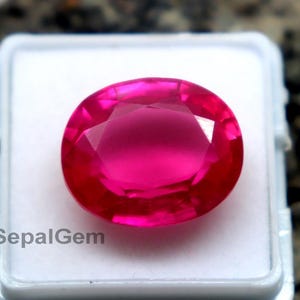 Può includere: Una vivace gemma rosa acceso, taglio ovale, esposta in una custodia di plastica bianca. La gemma è sfaccettata, catturando la luce e mostrando il suo colore brillante. Lo sfondo è una pietra strutturata, grigio scuro.