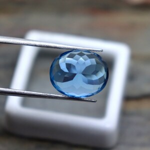 May include: A round, faceted, light blue gemstone held by tweezers. The gemstone is in a white square box.