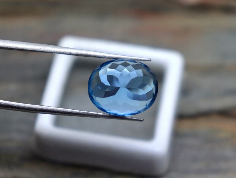 May include: A round, faceted, light blue gemstone held by tweezers. The gemstone is in a white square box.