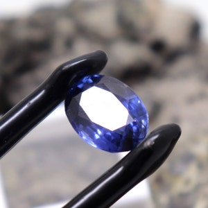 May include: A close-up of a blue oval-shaped gemstone held by a pair of black tweezers. The gemstone is faceted and has a bright, clear appearance.