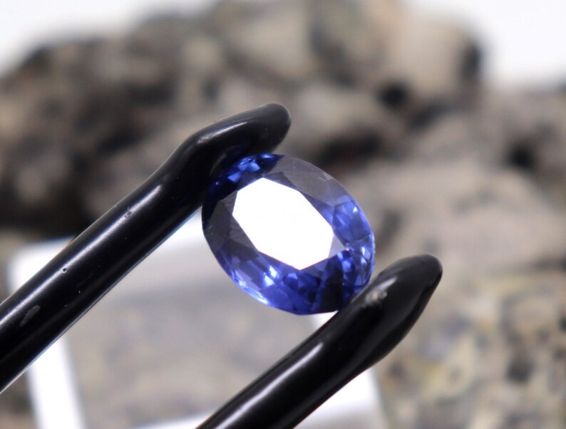 May include: A close-up of a blue oval-shaped gemstone held by a pair of black tweezers. The gemstone is faceted and has a bright, clear appearance.