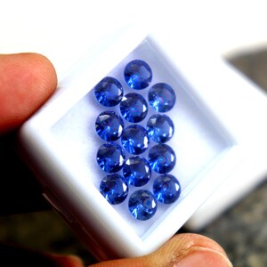 May include: A white box filled with round, blue gemstones. The box is being held by a hand.