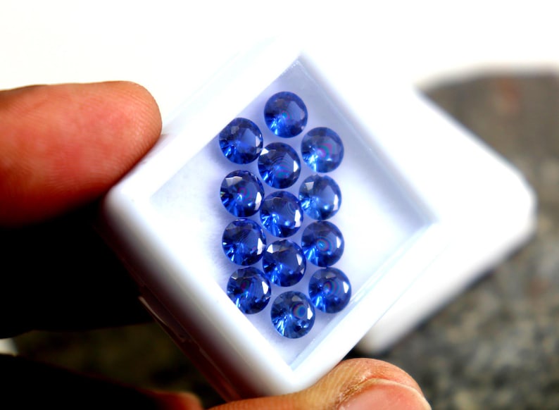 May include: A white box filled with round, blue gemstones. The box is being held by a hand.