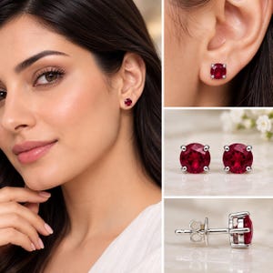 May include: A pair of round-cut red gemstone stud earrings set in a four-prong silver-toned metal setting. The earrings feature a faceted surface that reflects light, with a standard post and butterfly clutch backing mechanism for secure attachment.