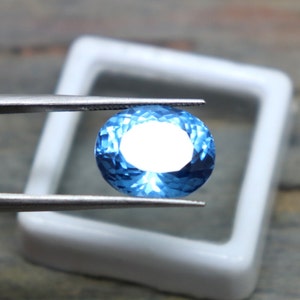 May include: A bright blue oval-shaped gemstone with a faceted cut. The gemstone is held by tweezers over a white background.