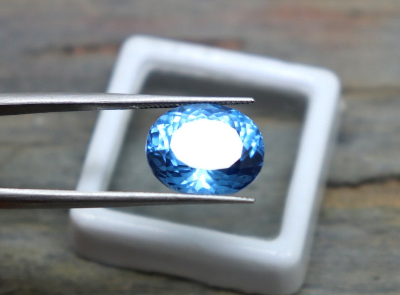 May include: A bright blue oval-shaped gemstone with a faceted cut. The gemstone is held by tweezers over a white background.