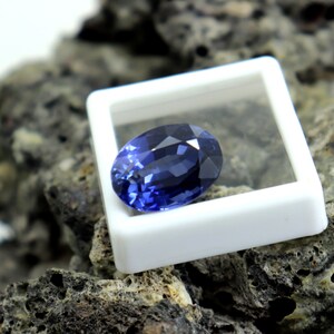 May include: A deep blue oval-shaped gemstone in a white display box. The gemstone is sitting on a rough, dark gray rock.