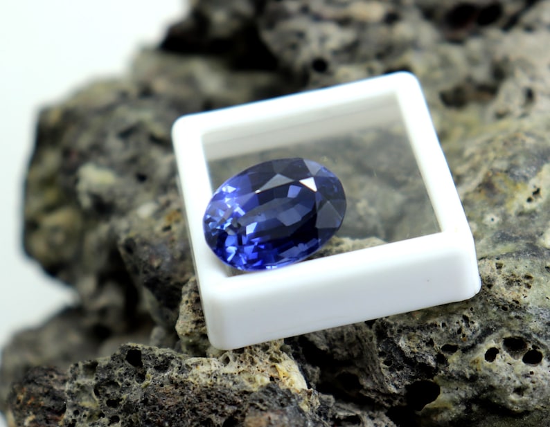 May include: A deep blue oval-shaped gemstone in a white display box. The gemstone is sitting on a rough, dark gray rock.