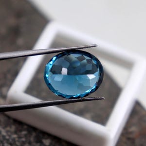 Puede incluir: Una gema azul facetada, de forma ovalada, es sostenida por unas pinzas sobre un fondo cuadrado blanco. La gema tiene un corte brillante, que refleja la luz. Las pinzas son plateadas y el fondo est&aacute; ligeramente desenfocado. La gema mide unos 2,5 cm de di&aacute;metro.