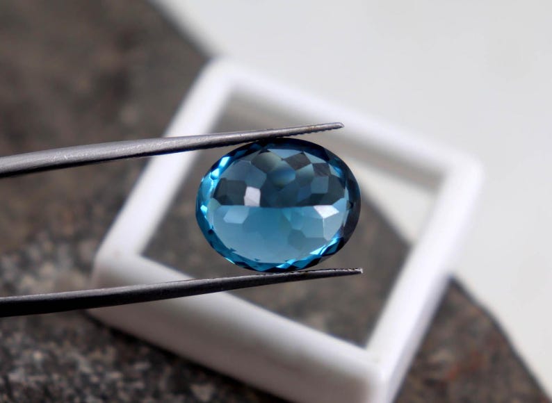 Puede incluir: Una gema azul facetada, de forma ovalada, es sostenida por unas pinzas sobre un fondo cuadrado blanco. La gema tiene un corte brillante, que refleja la luz. Las pinzas son plateadas y el fondo est&aacute; ligeramente desenfocado. La gema mide unos 2,5 cm de di&aacute;metro.
