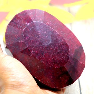 May include: A large, oval-shaped, faceted ruby gemstone with a deep red colour. The ruby has a polished finish and is held in a hand.