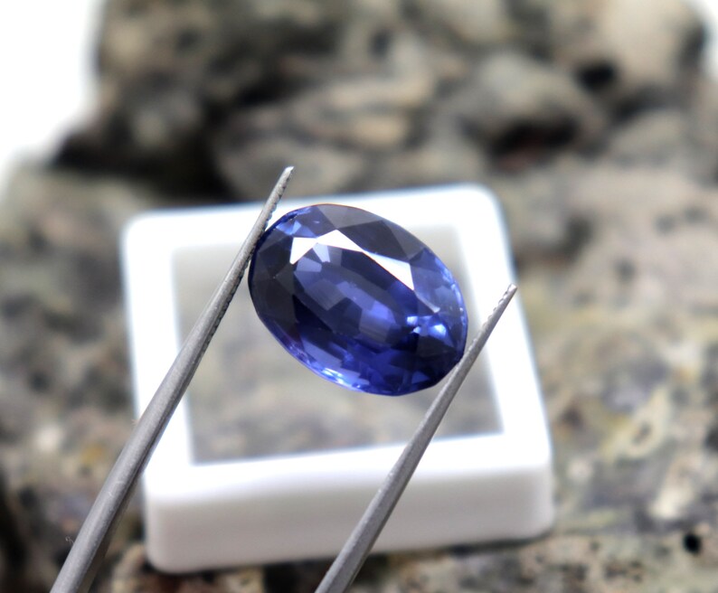 May include: A deep blue oval-shaped gemstone held by tweezers. The gemstone is faceted and has a high polish.