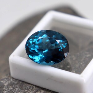 Puede incluir: Una gema facetada de forma ovalada, de color azul verdoso intenso. La gema se encuentra en un estuche cuadrado blanco, sobre una superficie gris texturizada. Las facetas de la gema reflejan la luz, creando un efecto brillante.