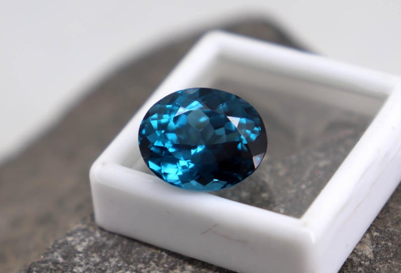 Puede incluir: Una gema facetada de forma ovalada, de color azul verdoso intenso. La gema se encuentra en un estuche cuadrado blanco, sobre una superficie gris texturizada. Las facetas de la gema reflejan la luz, creando un efecto brillante.