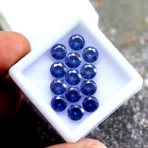May include: A close-up of a white box filled with 15 round, blue gemstones. The gemstones are arranged in a pyramid shape.