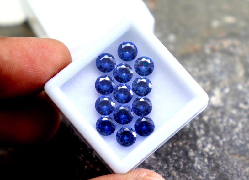 May include: A close-up of a white box filled with 15 round, blue gemstones. The gemstones are arranged in a pyramid shape.