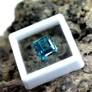 May include: A square-cut, teal-colored gemstone is displayed within a white plastic case. The gemstone has a brilliant cut, reflecting light. The case is set against a textured, gray-brown stone surface, highlighting the gem's color and clarity. This image showcases a precious stone.