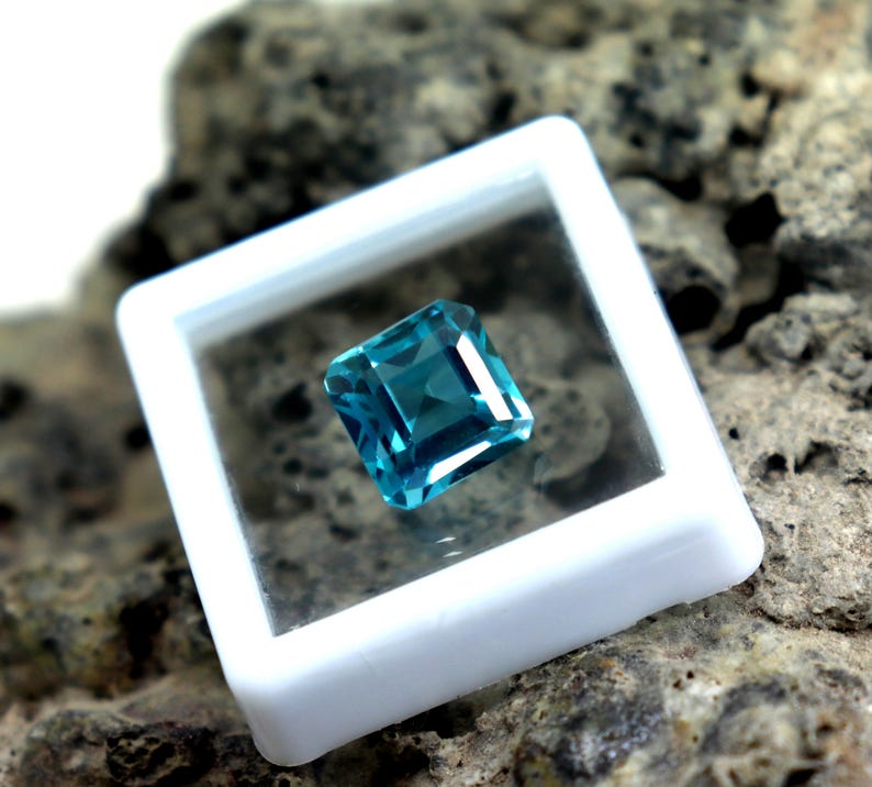May include: A square-cut, teal-colored gemstone is displayed within a white plastic case. The gemstone has a brilliant cut, reflecting light. The case is set against a textured, gray-brown stone surface, highlighting the gem's color and clarity. This image showcases a precious stone.
