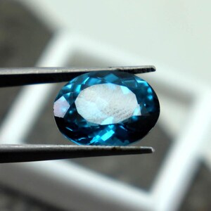Puede incluir: Una gema facetada de forma ovalada sostenida por unas pinzas. La gema es de color verde azulado intenso con una superficie brillante y reflectante. El fondo est&aacute; borroso, con una forma cuadrada blanca visible.