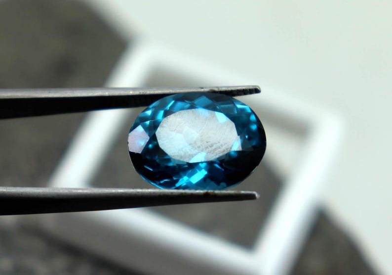 Puede incluir: Una gema facetada de forma ovalada sostenida por unas pinzas. La gema es de color verde azulado intenso con una superficie brillante y reflectante. El fondo est&aacute; borroso, con una forma cuadrada blanca visible.