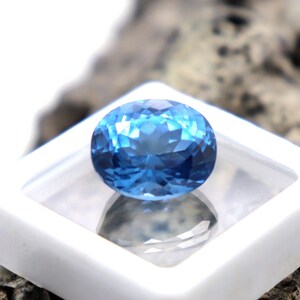 May include: A bright blue gemstone with a faceted oval shape, displayed in a white square box.