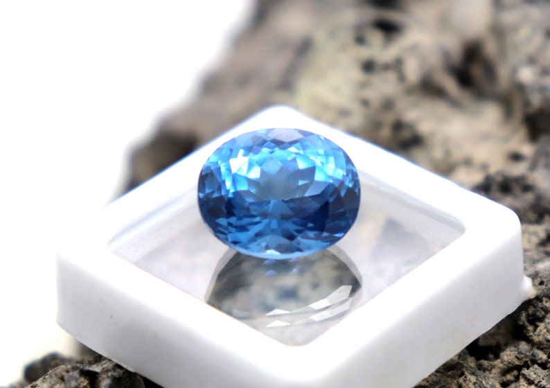 May include: A bright blue gemstone with a faceted oval shape, displayed in a white square box.
