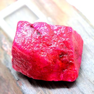 May include: A large, rough, red ruby gemstone. The ruby is a deep, rich red color and has a slightly textured surface.