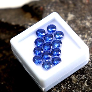 May include: A white box filled with round, blue gemstones. The box is sitting on a dark, textured surface.