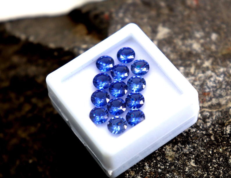 May include: A white box filled with round, blue gemstones. The box is sitting on a dark, textured surface.