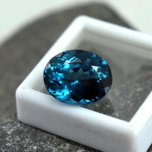 Puede incluir: Una gema ovalada facetada, de color azul verdoso intenso, dentro de una caja blanca. Las facetas brillantes capturan la luz. La caja se encuentra sobre una superficie gris texturizada, creando contraste.
