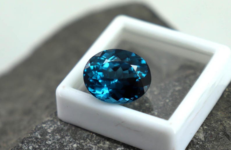 Puede incluir: Una gema ovalada facetada, de color azul verdoso intenso, dentro de una caja blanca. Las facetas brillantes capturan la luz. La caja se encuentra sobre una superficie gris texturizada, creando contraste.