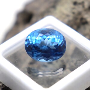 May include: A deep blue oval-shaped gemstone with a faceted cut. The gemstone is displayed in a white square box.