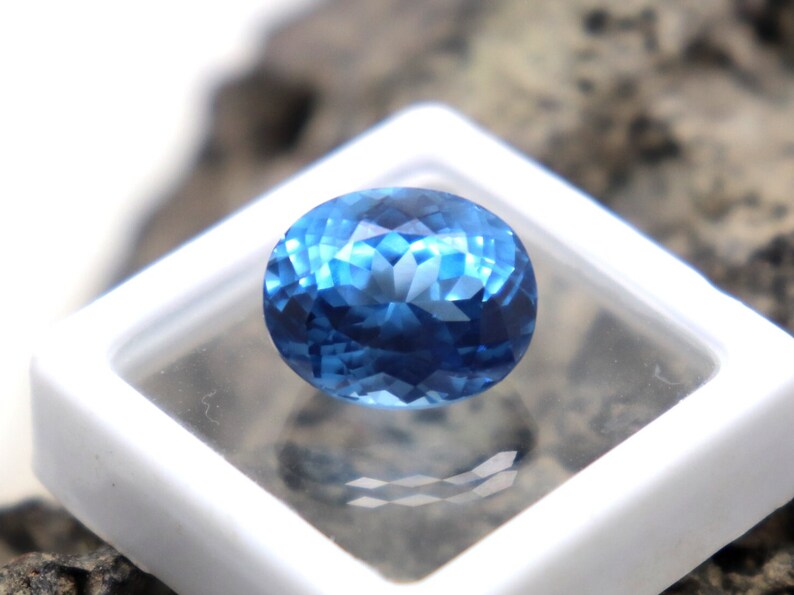 May include: A deep blue oval-shaped gemstone with a faceted cut. The gemstone is displayed in a white square box.