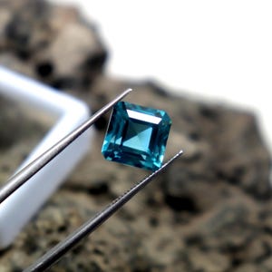 May include: A square-cut, teal gemstone is held by tweezers against a blurred background. The gem has a faceted surface, reflecting light. The tweezers are silver-colored, and a white object is partially visible in the background. The gemstone is the focal point.