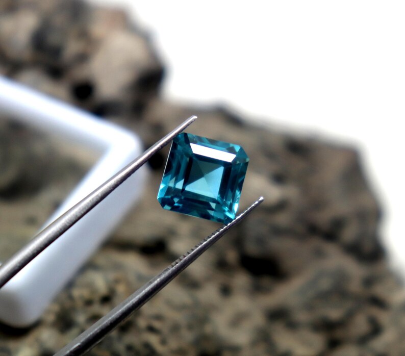 May include: A square-cut, teal gemstone is held by tweezers against a blurred background. The gem has a faceted surface, reflecting light. The tweezers are silver-colored, and a white object is partially visible in the background. The gemstone is the focal point.