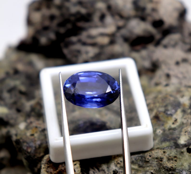 May include: A deep blue oval sapphire gemstone held by tweezers over a white box. The gemstone is a vibrant blue color and has a smooth, polished surface.