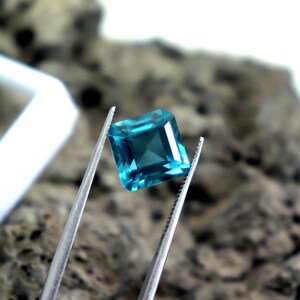 May include: A square-cut, teal gemstone is held by silver tweezers. The gem has a faceted surface that reflects light, creating a sparkling effect. The background is blurred, with a neutral color palette.
