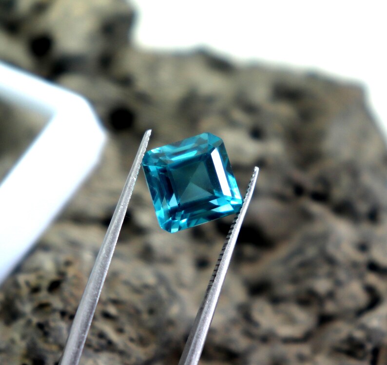 May include: A square-cut, teal gemstone is held by silver tweezers. The gem has a faceted surface that reflects light, creating a sparkling effect. The background is blurred, with a neutral color palette.