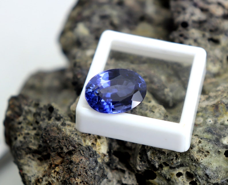 May include: A deep blue oval-shaped gemstone in a white display box. The gemstone is on a rough, dark gray rock surface.