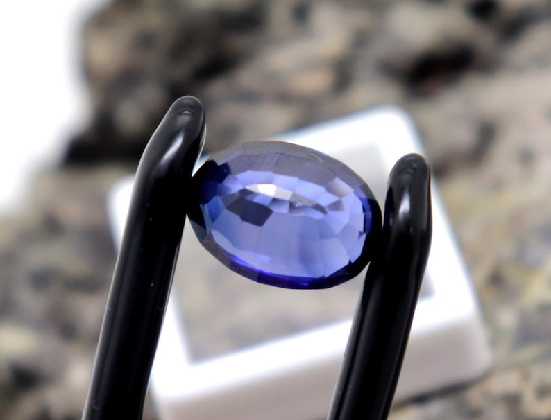 May include: A deep blue oval-shaped gemstone held in a pair of black tweezers.