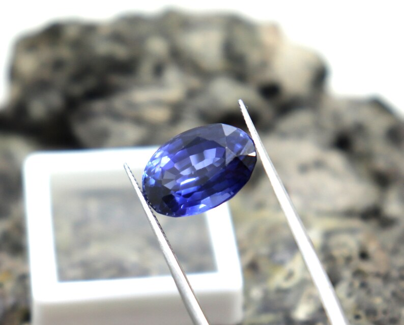 May include: A deep blue oval sapphire gemstone held by tweezers. The sapphire is faceted and has a brilliant shine.
