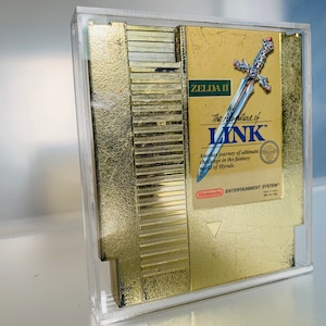 May include: A gold-colored Zelda II video game cartridge in a clear acrylic case. The cartridge label features the title "The Adventure of Link" with a sword illustration. The Nintendo Entertainment System logo is visible.